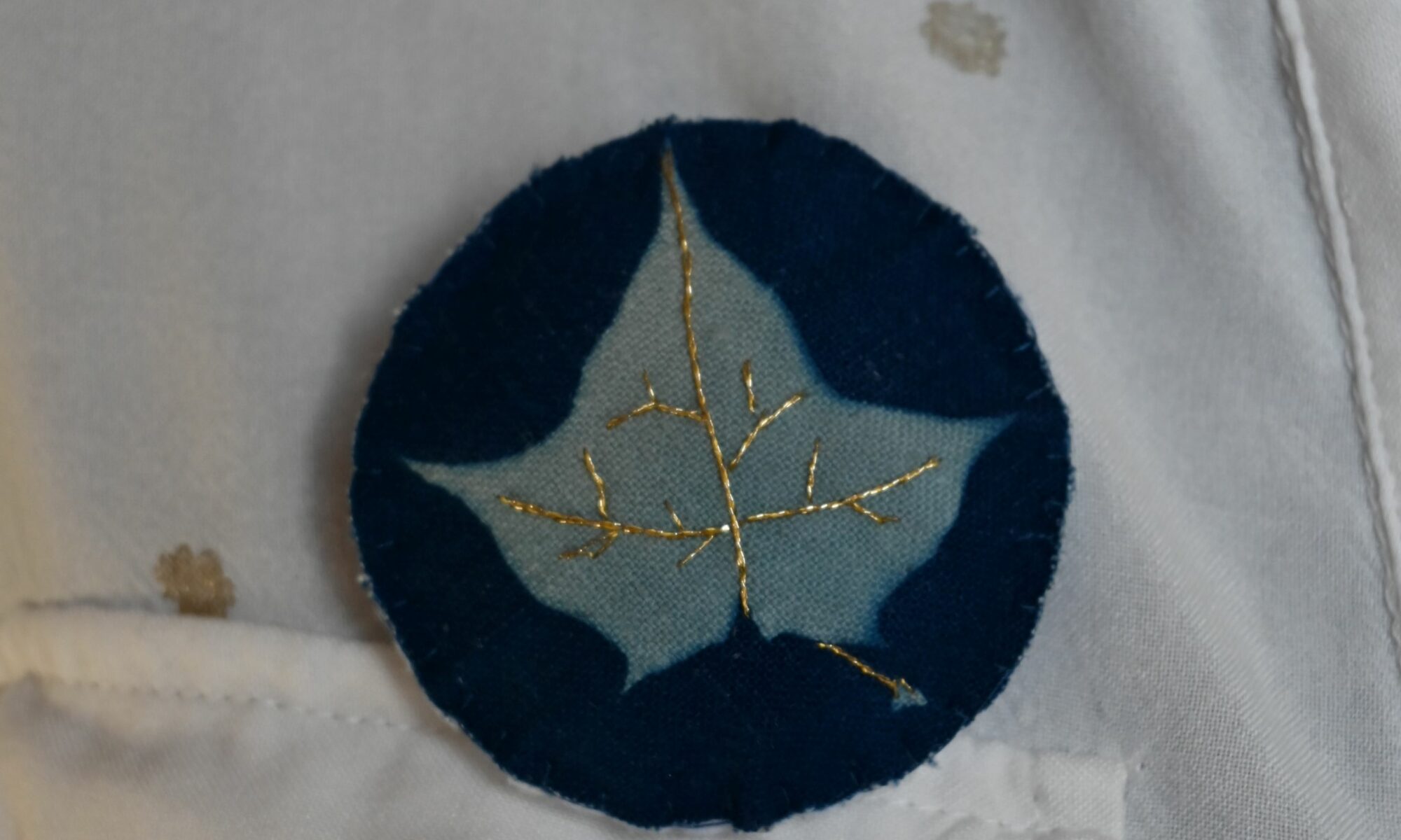 Une broche en tissu représentant une feuille d'érable révélé par le cyanotype et brodé au fil d'or