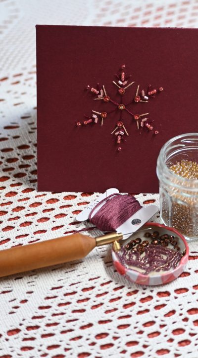 une carte brodée, de perles dans les tons rose foncé, perles, tubes, paillettes et fil doré, on voit un poinçon, du fil métallisé et des perles dorées au premier plan